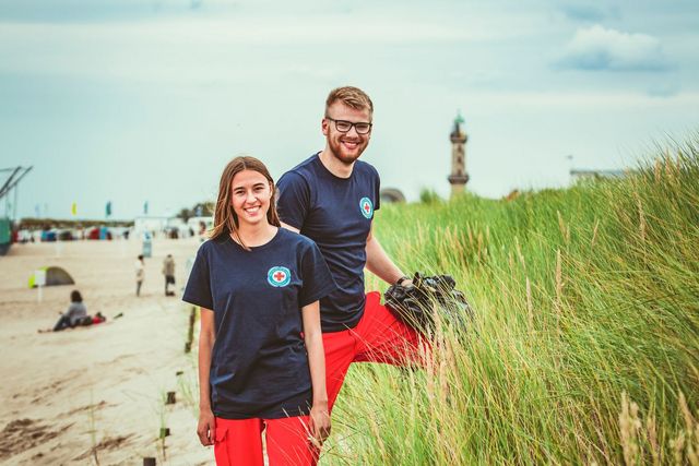 Zwei ehrenamtlich Aktive der Wasserwacht stehen an einer Düne und lächeln in die Kamera